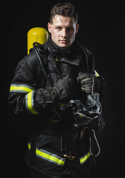 Modisches Porträt eines jungen Mannes in Feuerwehrausrüstung auf schwarzem Studiohintergrund. Feuerwehrmann in dunkler Uniform - Foto, Bild
