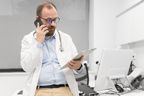 Steteskoplu bir erkek doktor masada oturuyor, cep telefonuyla konuşuyor ve dijital tablete bakıyor, bir hastanın teşhisini tartışıyor.. - Fotoğraf, Görsel