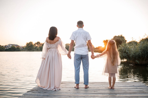 妊娠中の妻と娘を持つ夫が川岸に腰を下ろしており、夏の晴れた日の日没時に桟橋に立っている。男と女が手を握って抱き合ってる。幸せな家族 - 写真・画像