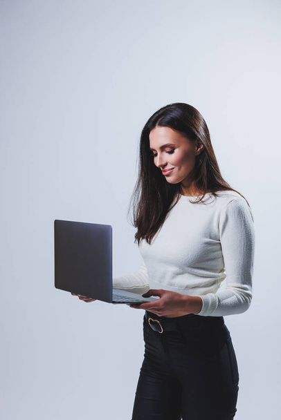 Young woman manager in a white shirt holds a portable laptop in her hands while standing on a gray background - Foto, immagini