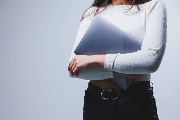 Молодая женщина менеджер в белой рубашке держит портативный ноутбук в руках, стоя на сером фоне - Фото, изображение