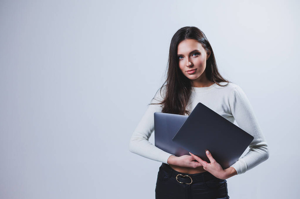 Young woman manager in a white shirt holds a portable laptop in her hands while standing on a gray background - Foto, Imagen