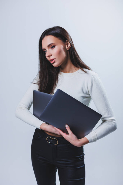 Young woman manager in a white shirt holds a portable laptop in her hands while standing on a gray background - Φωτογραφία, εικόνα