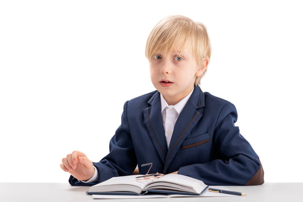Blond boy of 7-8 years old in jacket and white shirt sits at table in front of open notebook. Small child imitates the director - Photo, Image