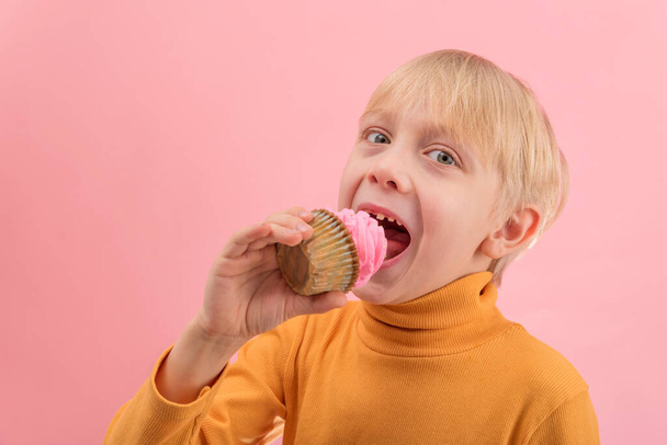 Fair-haired boy eats cupcake with pink icing. Child looks at the camera and wants to bite off piece of muffin - Photo, Image