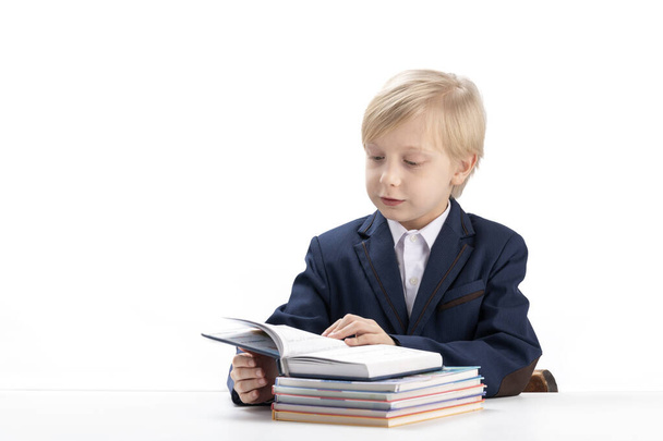 Blonde schoolboy sits at table and flips through textbook. Boy in school uniform reads textbook. Bright student - Fotografie, Obrázek
