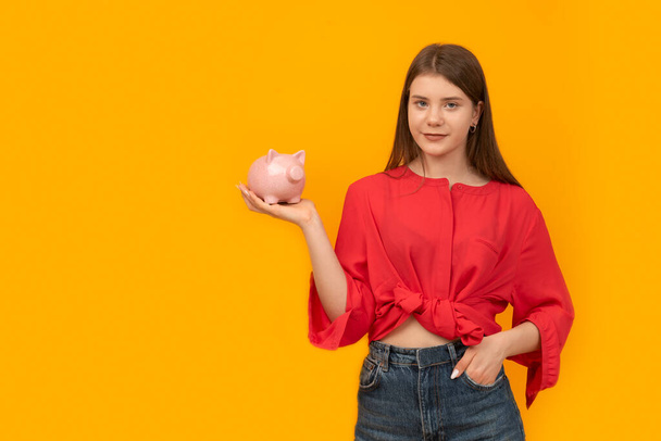 Teenage girl holds piggy bank in her hands on yellow background. Financial literacy. Investment. Money box - Foto, Imagen