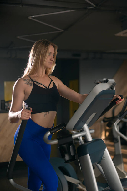 Luxurious young woman with blonde hair is engaged in gym on an orbitrek elliptical trainer. Vertical frame - Foto, imagen