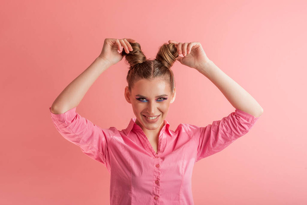 Radosna młoda kobieta w różowej koszuli dopasowuje włosy. Dziewczyna z dwiema fryzury bułki na różowym tle - Zdjęcie, obraz