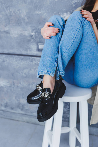 Na židli ve studiu sedí mladá žena s štíhlýma krásnýma nohama v módních černých kožených botách. Dámské stylové mokasíny, detailní záběr. Jarní kolekce bot - Fotografie, Obrázek