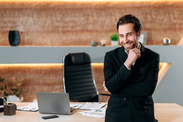 Портрет элегантного кавказского успешного бизнесмена в формальном стильном костюме, предпринимателя, генерального директора компании, стоящего в своем современном офисе рядом с рабочим столом, смотрящего в камеру, радостно улыбающегося - Фото, изображение