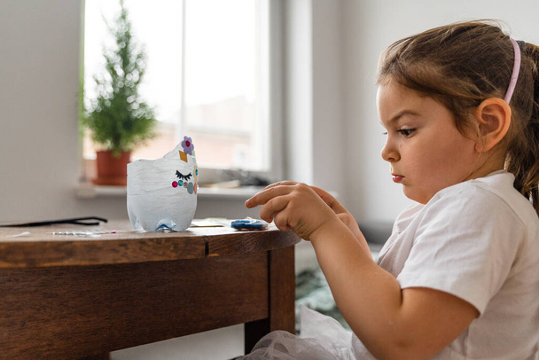 Piękna biała przedszkolanka bawiąca się w domu i robiąca rzemiosło z plastikowej butelki. Recykling zabawek i kreatywność  - Zdjęcie, obraz