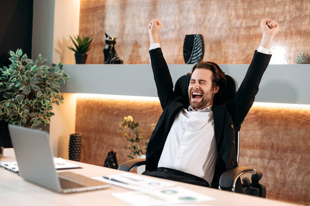 Success, joy, victory concept. Caucasian happy businessman in a formal stylish suit, sit in his modern office, rejoicing at a great deal, financial profit, successful project, gesturing with his hands - Photo, Image
