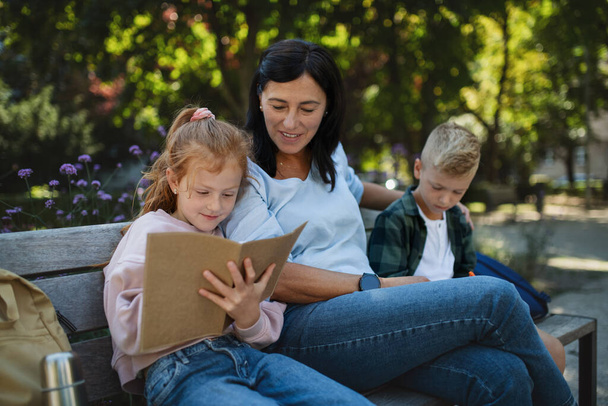 Happy senior woman with grandchildren sitting on bench and helping with homework outdoors in park. - Фото, изображение