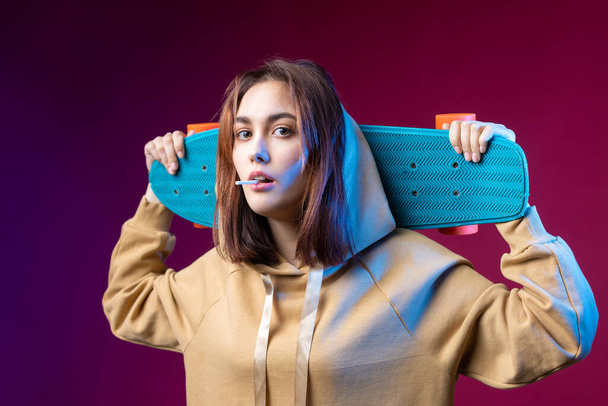 Νεαρή όμορφη μοντέρνα hipster κορίτσι ντυμένη με κουκούλα κρατά ένα skateboard στα χέρια της σε ένα μωβ φόντο στούντιο. Τρώει ένα γλειφιτζούρι. - Φωτογραφία, εικόνα