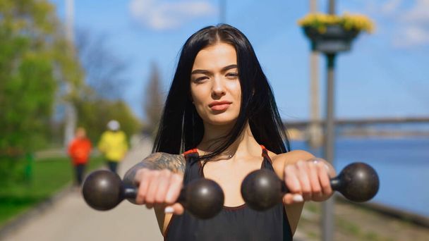 Serious brunette woman holds two old dumbbells during sunny day and looks at you - Foto, imagen