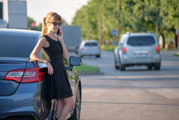Młoda kobieta kierowca stojąca w pobliżu samochodu rozmawia przez telefon komórkowy na ulicy miasta w lecie - Zdjęcie, obraz