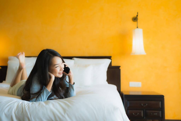 寝室のインテリアでコーヒーカップとベッドの上で携帯電話を使用して肖像画の美しい若いアジアの女性 - 写真・画像