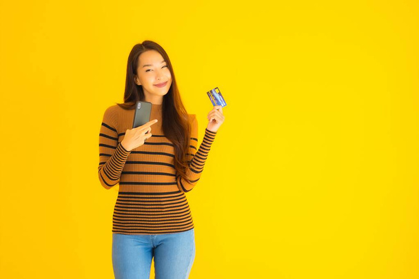 肖像画の美しい若いアジアの女性は黄色の隔離された背景にオンラインショッピングのためのクレジットカードでスマートフォンや携帯電話を使用します - 写真・画像