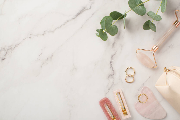 Skincare beauty concept. Top view photo of pink barrettes rose quartz roller gua sha gold rings cosmetics bag and eucalyptus on white marble background with copyspace - Photo, Image