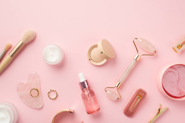 Top view photo of makeup brushes rose quartz roller gua sha pink eye patches glass transparent bottle cream jars eyeshadow barrettes lip gloss gold rings and wristlet on isolated pink background - Photo, Image