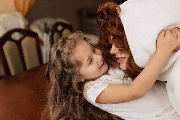 Close up indoor portrait of red haired woman in hoodie is hugging and spend time with her adorable little daughter at home. High quality photo - 写真・画像