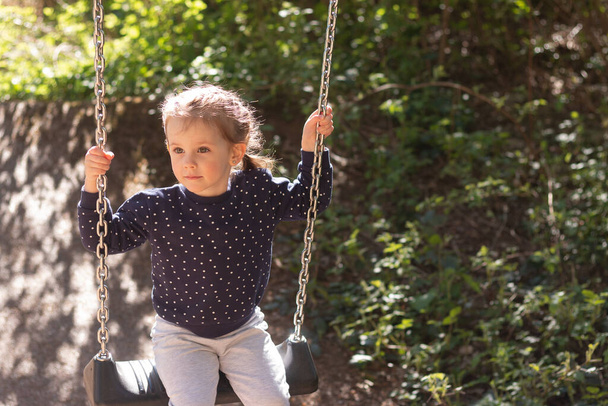 Μικρό όμορφο κορίτσι χαμογελά κούνιες σε μια κούνια στο πάρκο με φόντο το θολό φύλλωμα - Φωτογραφία, εικόνα