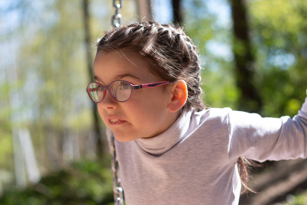 Little cute girl in glasses smiling rides on a swing at the playground in the park - Foto, immagini