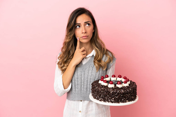 Jeune femme tenant gâteau d'anniversaire sur fond rose isolé ayant des doutes tout en levant les yeux - Photo, image