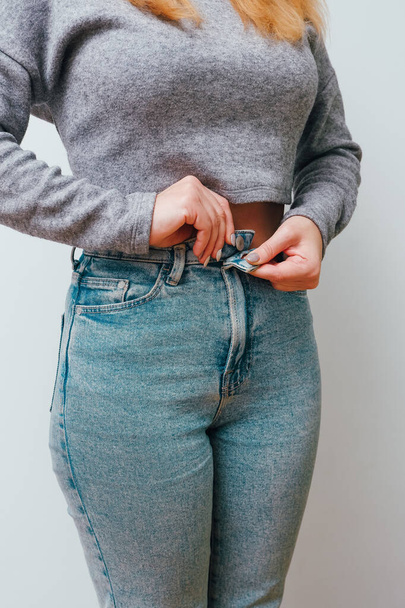A slender woman fastens a button of her blue jeans. Weight ungain woman getting dressed wearing jeans. Concept of excessive weight - Фото, изображение