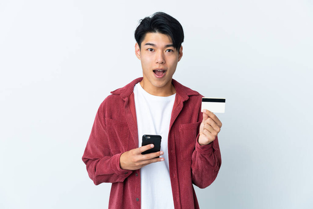 Young Chinese man isolated on white background buying with the mobile and holding a credit card with surprised expression - 写真・画像