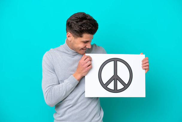 Young handsome caucasian man isolated on blue bakcground holding a placard with peace symbol - Foto, Imagem