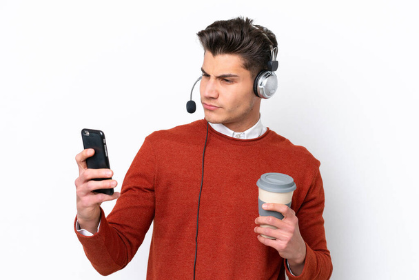 Telemarketer hombre caucásico trabajando con un auricular aislado sobre fondo blanco sosteniendo café para llevar y un móvil - Foto, imagen