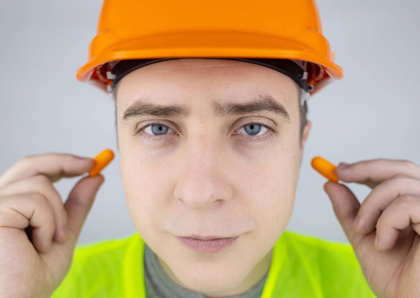 A worker in a hard hat and green vest puts on ear plugs. Industrial safety. Industrial noise protection. Compliance with the rules of conduct at loud work. Orange ear plugs - Фото, изображение