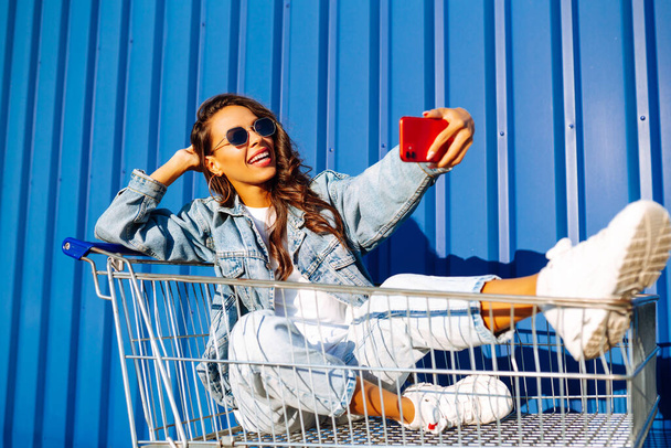 Selfie time. Young woman  having fun on shopping trolleys. Consumerism, sale, discounts, lifestyle concept. - Fotó, kép