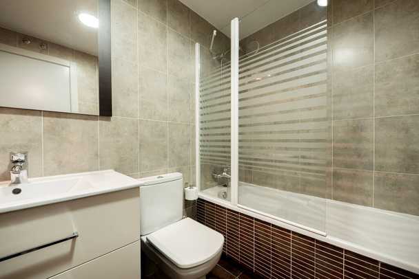 Ванная комната с раковиной из белой смолы на деревянном шкафу с ящиками, белыми туалетами, зеркалом на стене, стеклянной душевой кабиной и коричневой плиткой - Фото, изображение