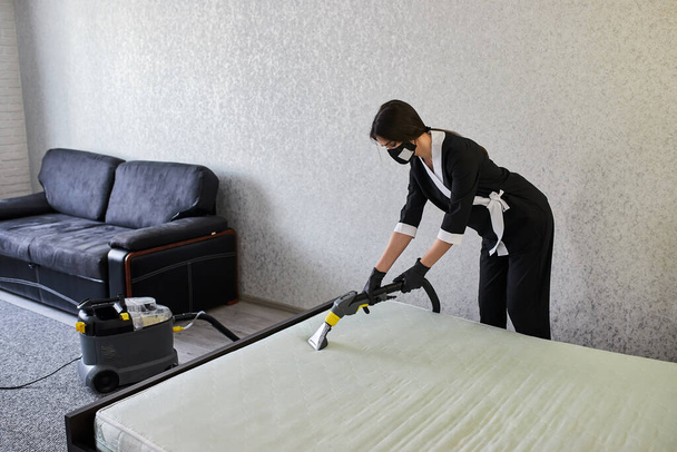 Siivouspalvelun työntekijä poistaa likaa huonekaluista litteässä ammattikäyttöön tarkoitetuilla laitteilla. Nainen kotiäiti puhdistaa patja sängyllä pesu pölynimurilla - Valokuva, kuva