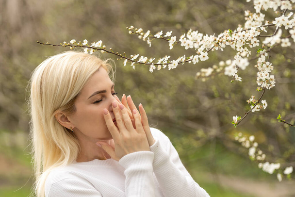Woman allergic suffering from seasonal allergy at spring, posing in blossoming garden at springtime. Young woman sneezing in front of blooming tree. Spring allergy concept - Photo, Image