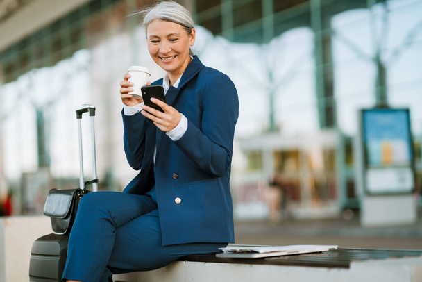 Grey asian woman using cellphone and drinking coffee by airport outdoors - Fotoğraf, Görsel