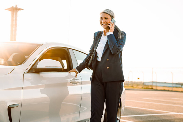 Mature asian businesswoman smiling and talking on cellphone while standing by car - Foto, immagini