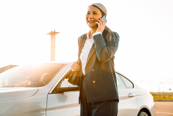 Mature asian businesswoman smiling and talking on cellphone while standing by car - Photo, image