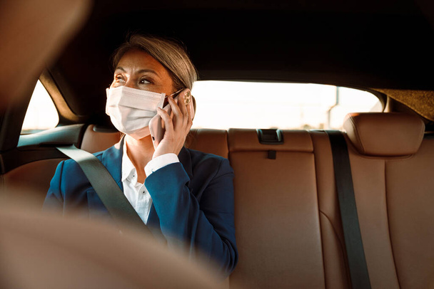 Mature asian businesswoman wearing face mask talking on mobile phone in car - Фото, изображение