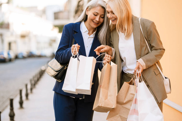 Mature multiracial women talking and looking in shopping bags while walking outdoors - Photo, image