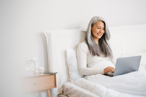 Grey asian woman smiling and using laptop while sitting in bed after sleep at home - Fotografie, Obrázek