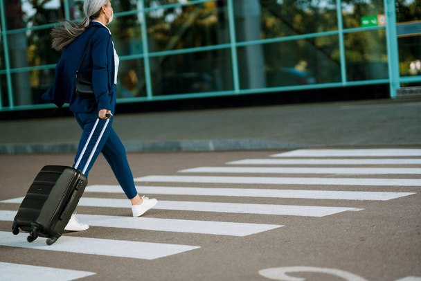 Grey woman in face mask walking with suitcase on zebra crossing outdoors - Фото, изображение