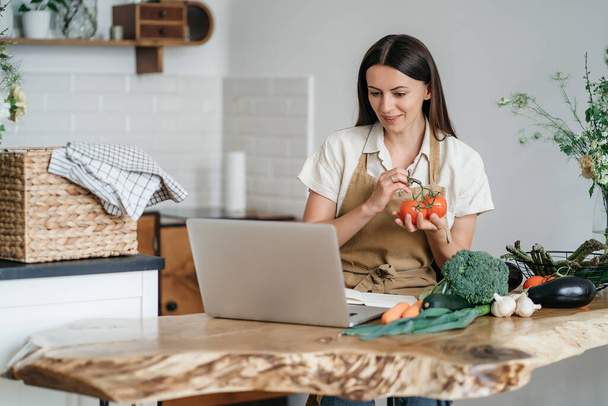 Una giovane donna sta usando un computer portatile in cucina per preparare verdure vegane sane. Corsi di cucina online - Foto, immagini