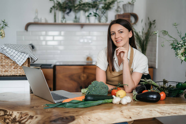 Genç bir kadın mutfakta dizüstü bilgisayar kullanıyor ve sağlıklı vegan sebzeler hazırlıyor. İnternette aşçılık eğitimi - Fotoğraf, Görsel
