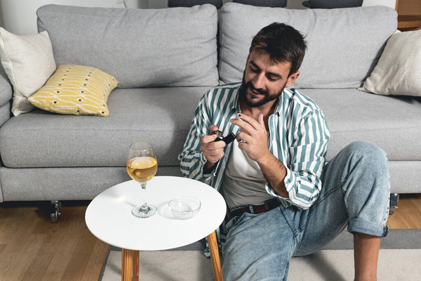 Young carefree man sitting on the floor in his home lighting a cigarette to smoking and drinks wine he has decided not to be unhappy and depressed anymore because life is only one and he wants to live - Fotoğraf, Görsel