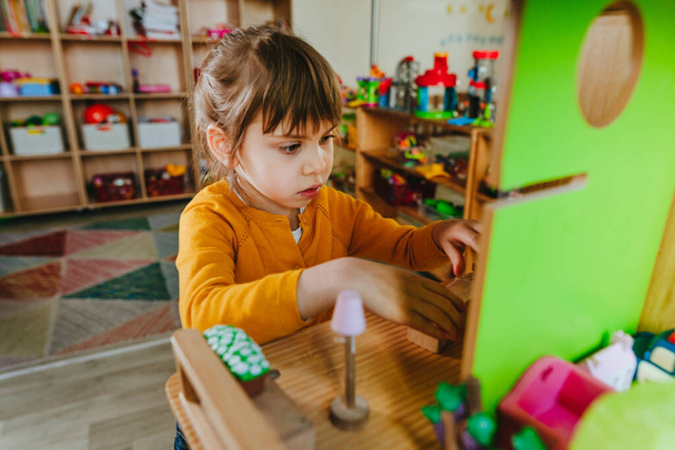 Little girl playing with wooden house with toy furniture at kindergarten or childcare centre. Early development with natural material toys. Montessori education concept. - Foto, Imagem