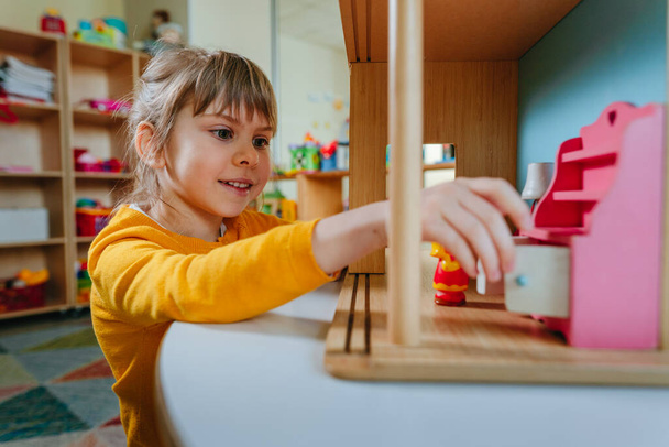 Kislány játszik faház játékbútorokkal óvoda vagy gyermekgondozási központ. Korai fejlesztés természetes anyagi játékokkal. Montessori oktatási koncepció. - Fotó, kép